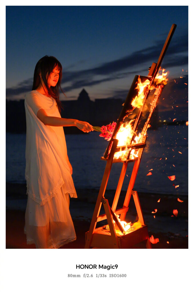 Photo d’exemple officielle du Honor Magic 9 prise de nuit au téléobjectif de 80 mm : une femme en robe blanche près d’un chevalet en flammes sur une plage, au crépuscule